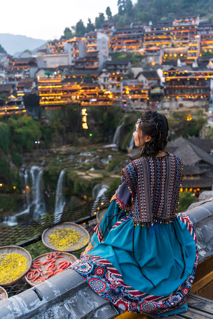 Una mujer vestida con ropa tradicional contempla la cascada del casco antiguo de Furong y las casas sobre pilotes iluminadas por la luz.