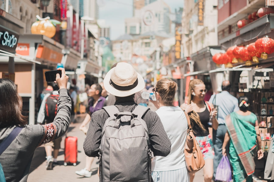 Turistas con mochilas explorando una concurrida calle del mercado de Chinatown, con sus faroles rojos y tiendas.